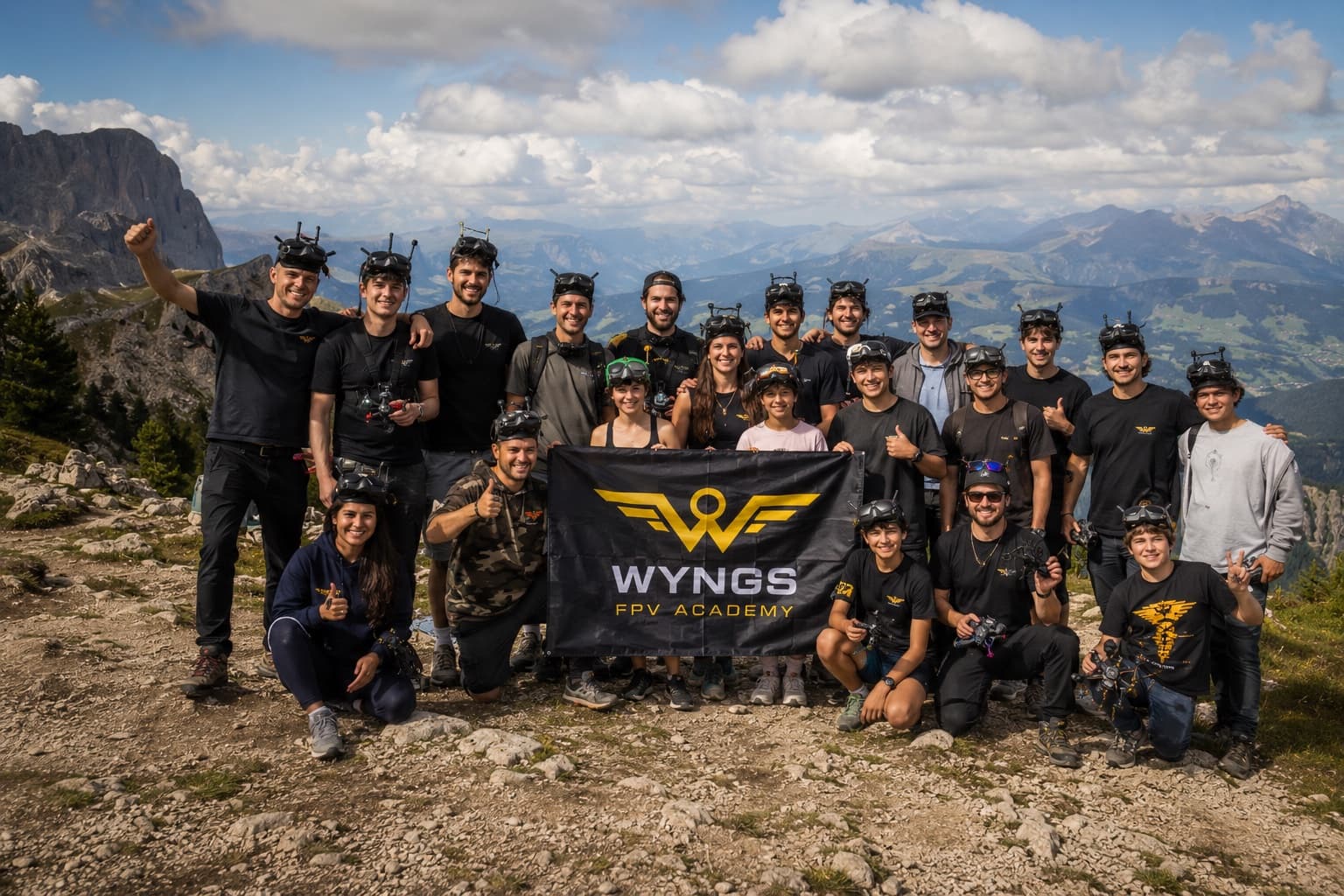 Around twenty WYNGS pilots on a mountain summit, goggles pushed up on their heads and quads in hand, crowded around a WYNGS banner with the valley behind them.