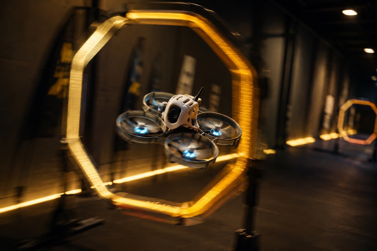 A WYNGS cinewhoop threading an illuminated gate at speed in a dark indoor hallway, motion blur streaking the propeller ducts and the gate's ring.