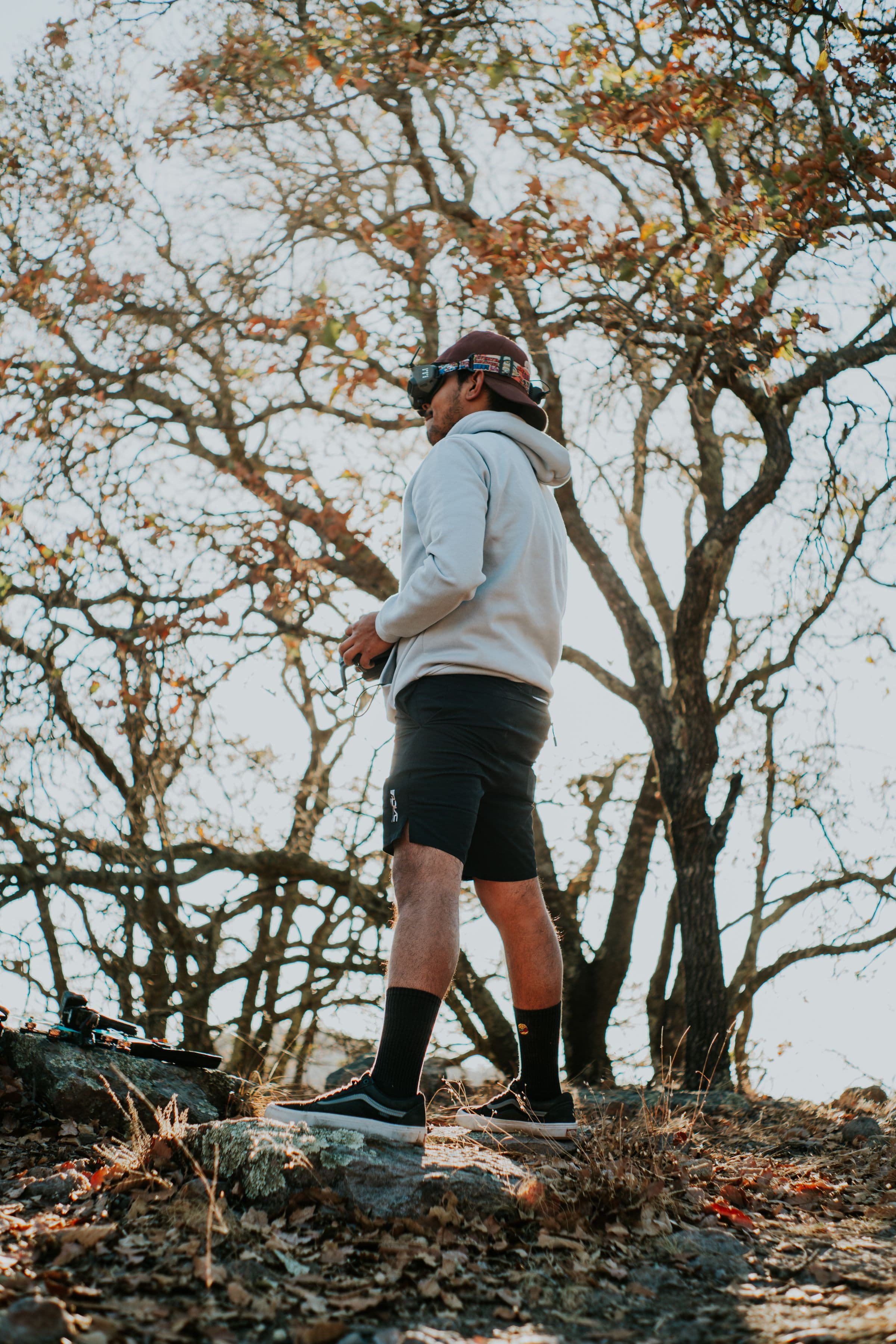 An adult wearing FPV goggles pauses in a quiet autumn woodland with a compact freestyle quadcopter resting on the rocks