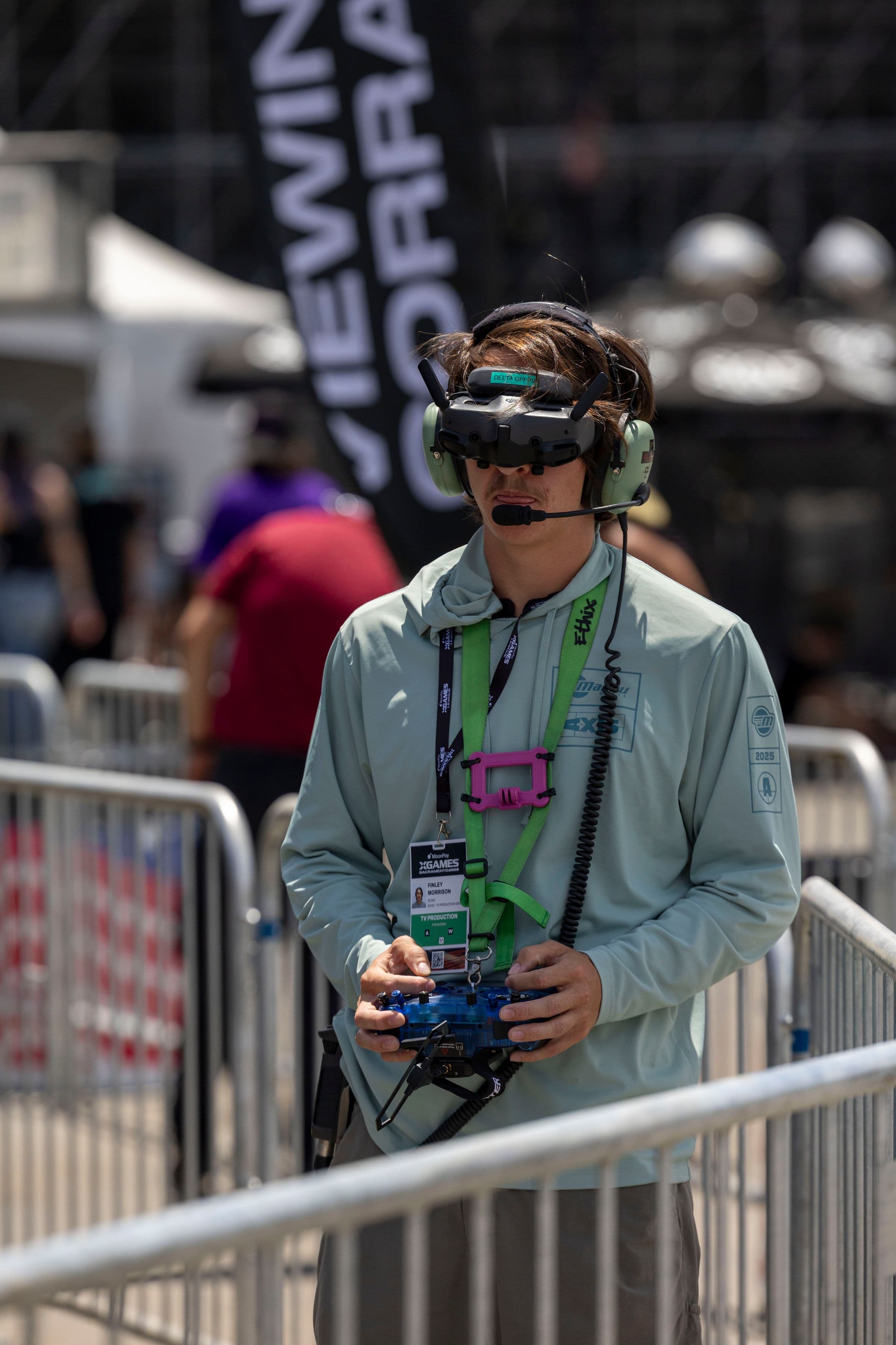 An FPV pilot wearing racing goggles adjusts a compact transmitter with exposed gimbal sticks at an outdoor flight event