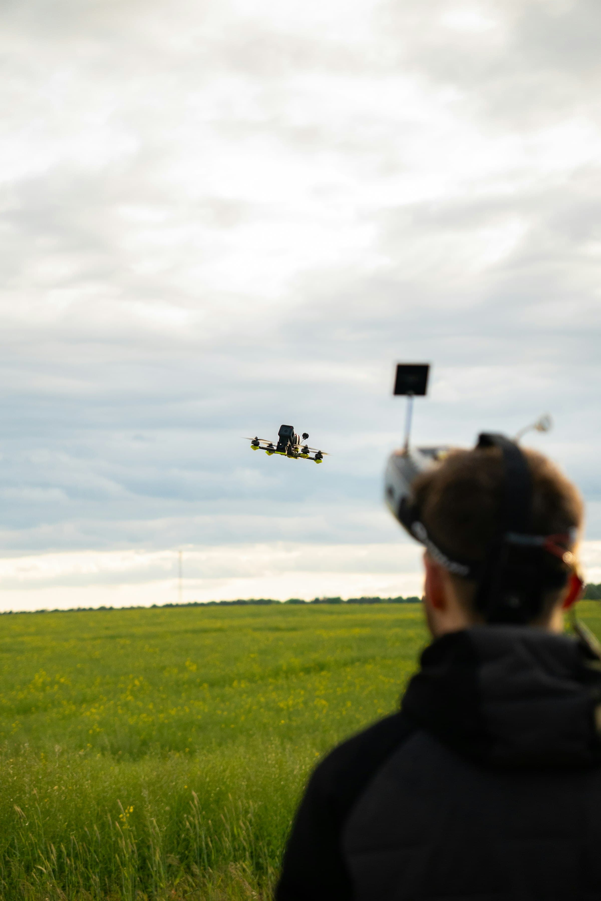 A pilot with a transmitter flying a small FPV quadcopter low over an open green field